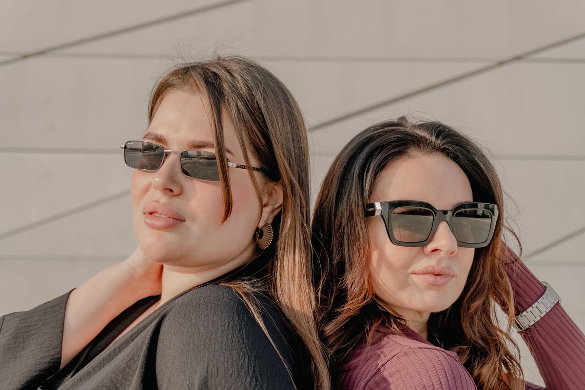 Two women in narrow and square sunglasses against a pale wall